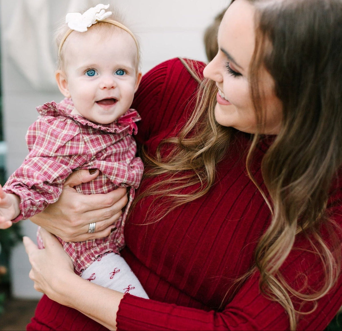 A woman in a red ribbed knit dress holds a baby wearing a matching red and pink plaid outfit with white socks. 