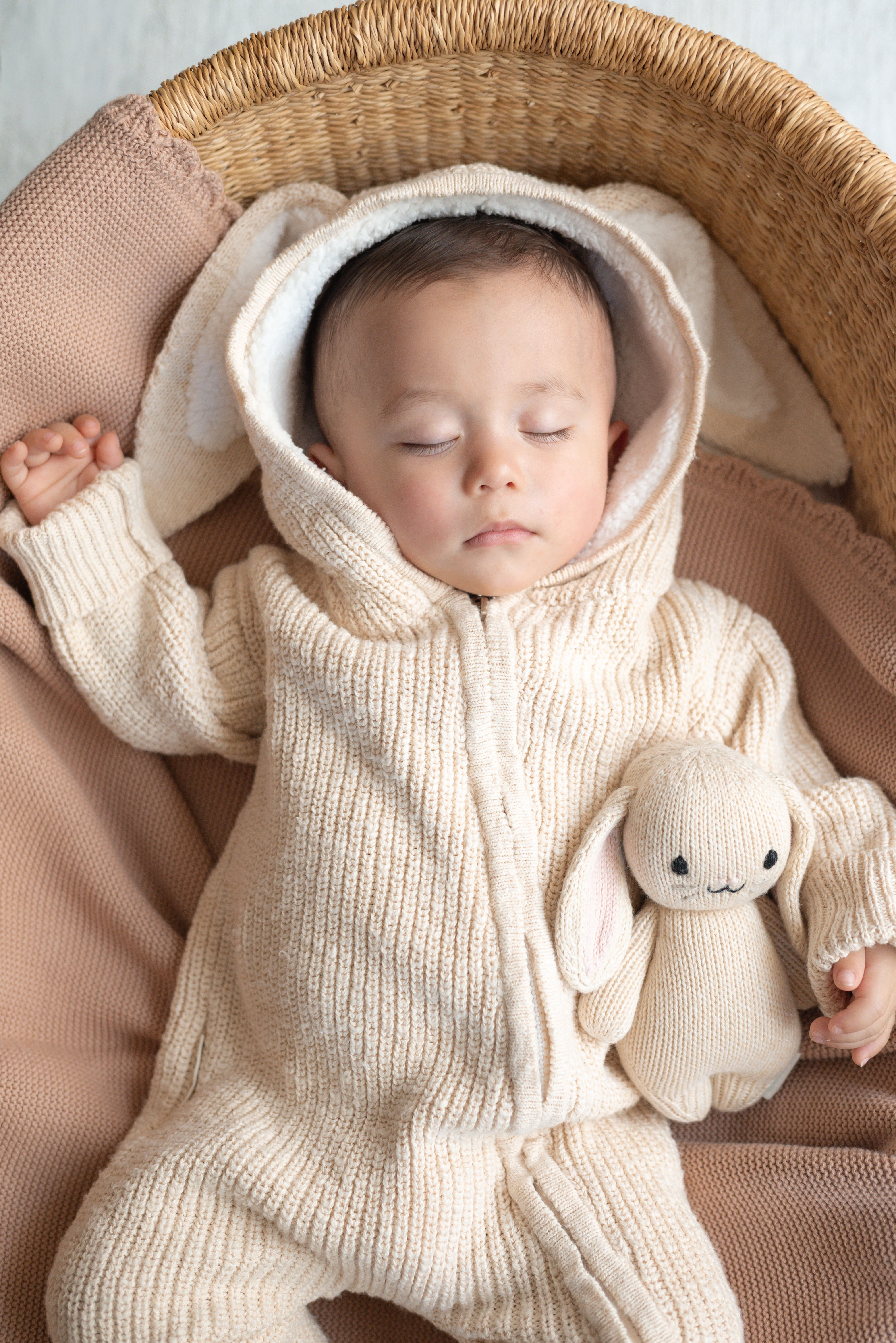 Baby in a beige knitted outfit with a bunny hat and toy, lying in a woven basket.