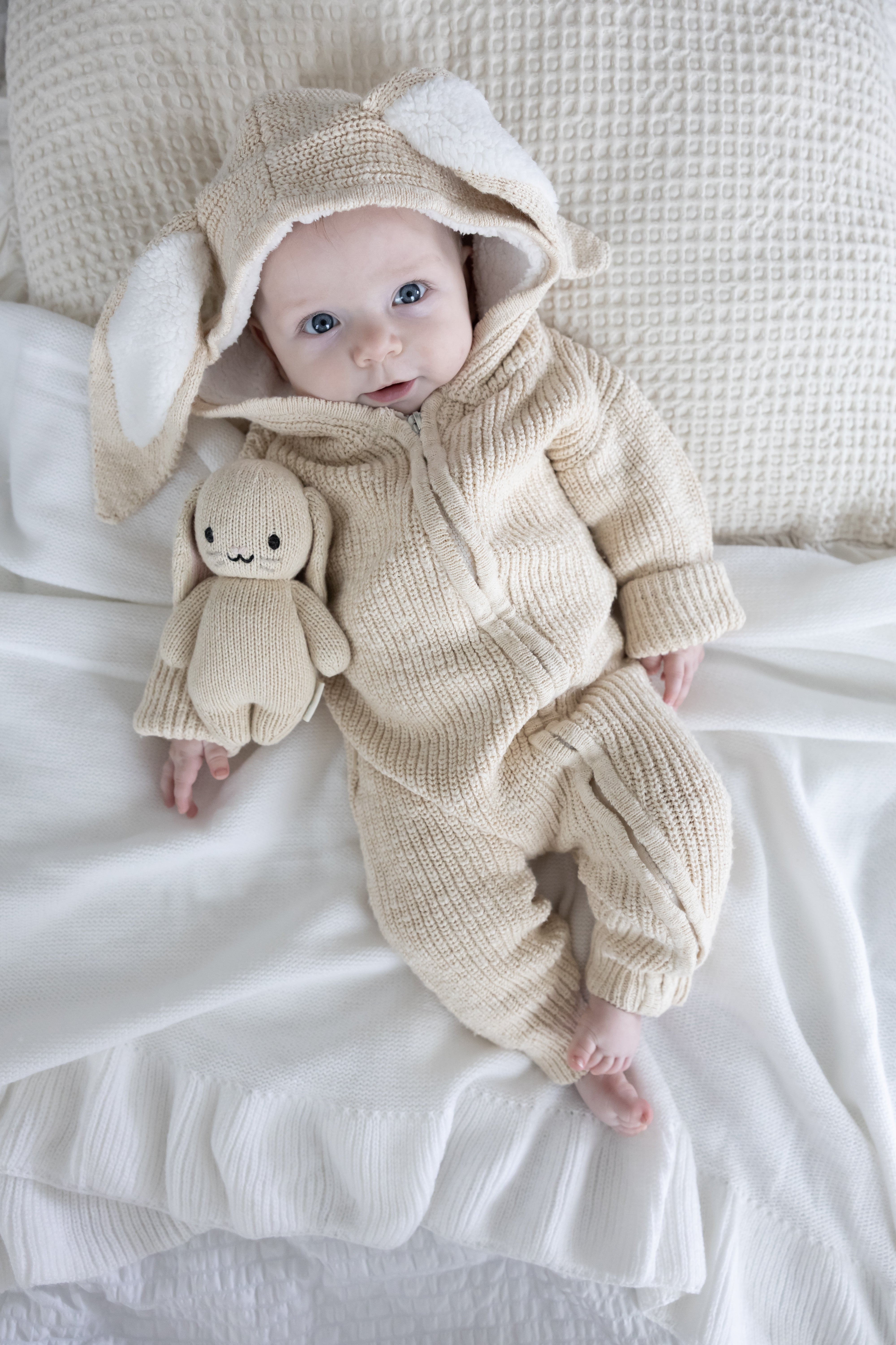 Baby in a beige bunny knit romper lying on a white blanket