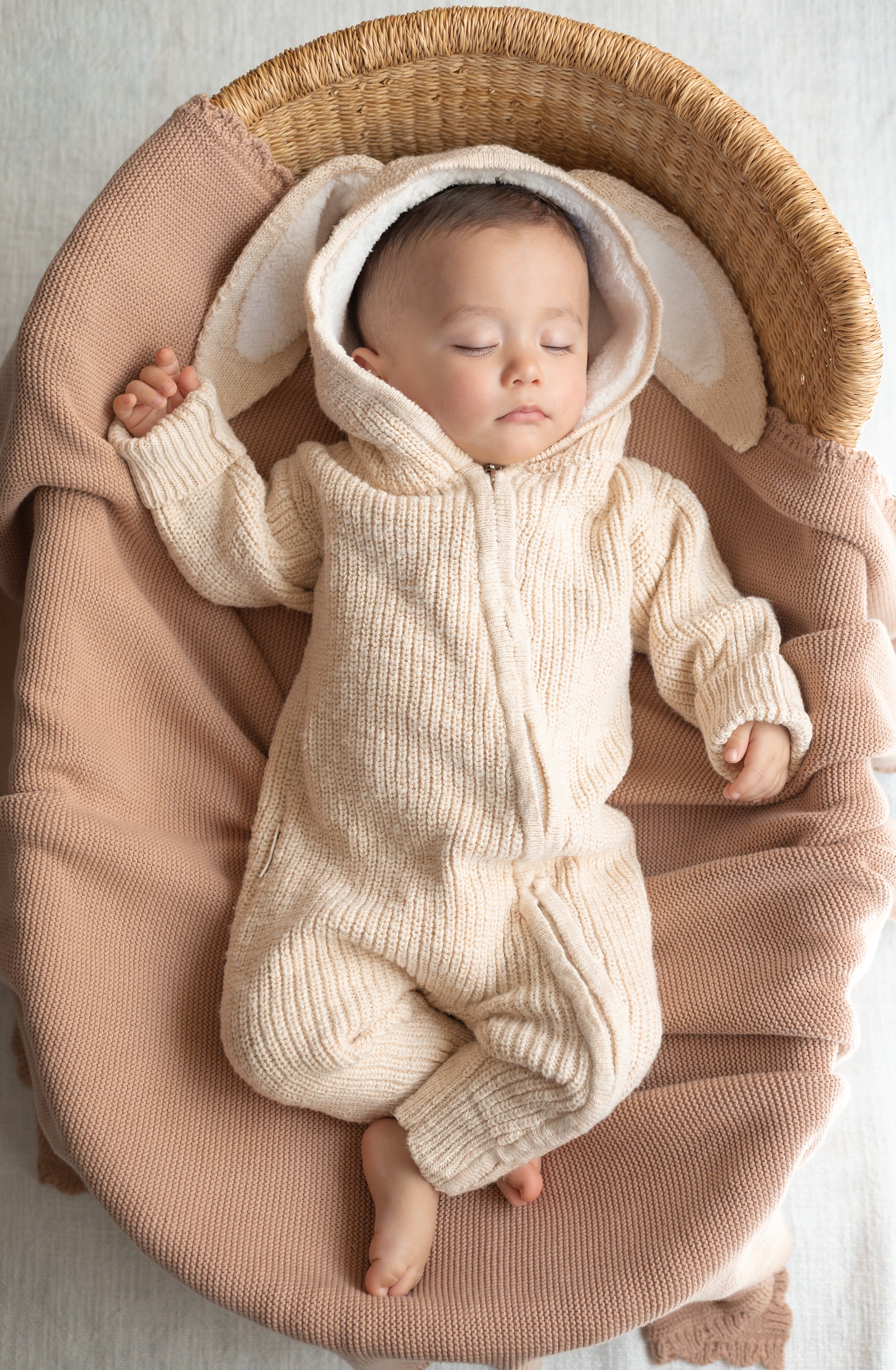 Baby in a cream-colored knitted outfit with bunny ears lying in a woven basket.