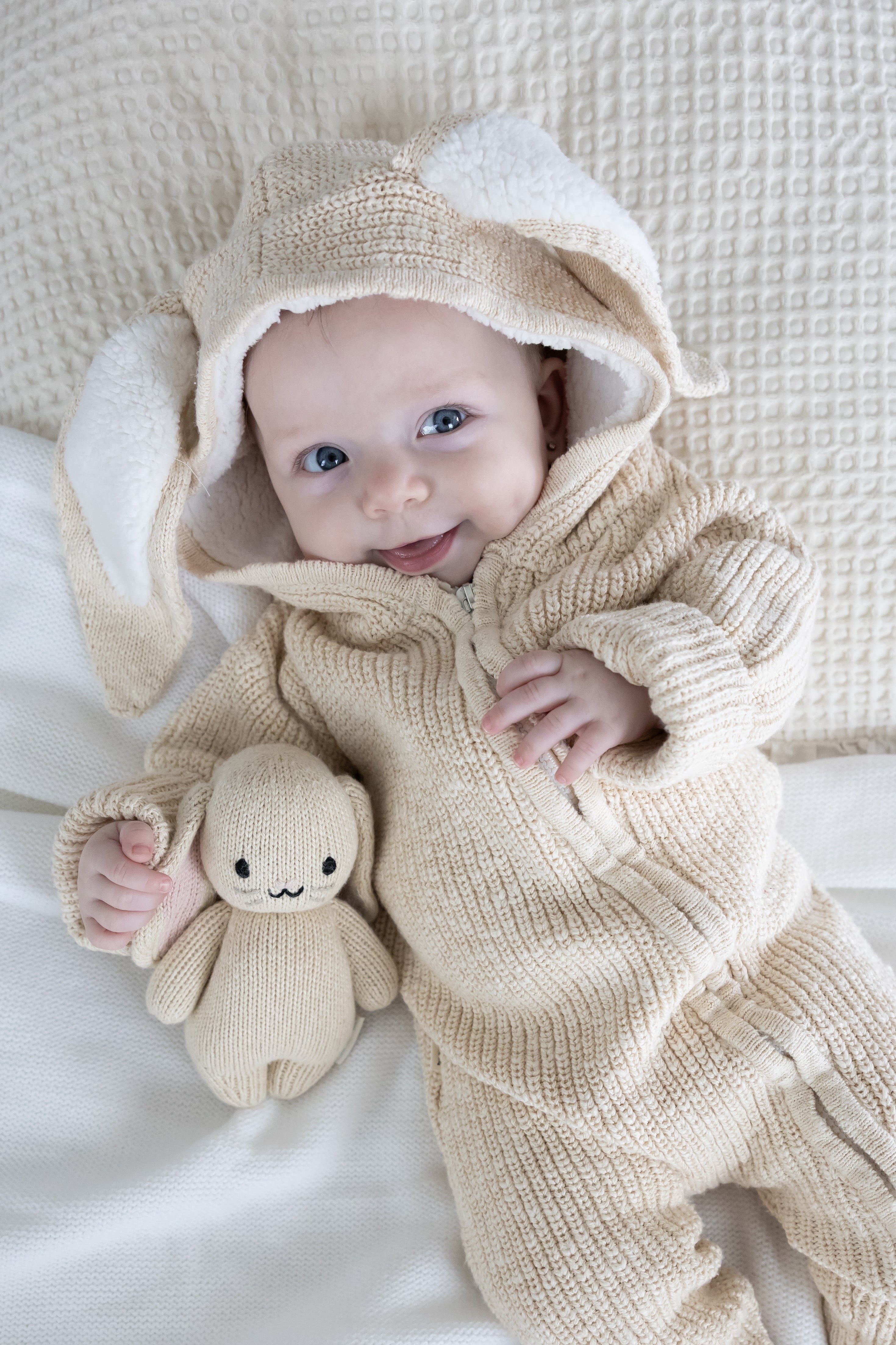 Baby in a beige knitted outfit with bunny ears holding a plush toy on a white blanket.