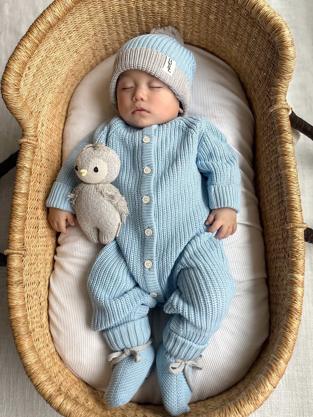 A sleeping newborn wearing the Coastal Blue Keepsake Knit Romper, matching Knit Pom Beanie, and Knit Tie Booties. The baby is tucked into a woven bassinet next to a small grey knit penguin plush toy.